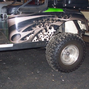 Könnte beinhalten: Ein EZGO Golfwagen-Seitenteil mit einer individuellen Airbrush-Grafik aus mehreren Totenköpfen mit schwarzen und grauen flammenartigen Akzenten. Das Fahrzeug verfügt über einen schwarzen Sitz, eine grüne Zierleiste und einen grobstolligen Geländereifen.