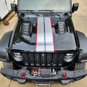 May include: Black Jeep with a silver and red racing stripe down the bonnet. The front bumper has a black grill and a winch with the word "BADLAND" on it. The tyres are black and have a rugged tread.