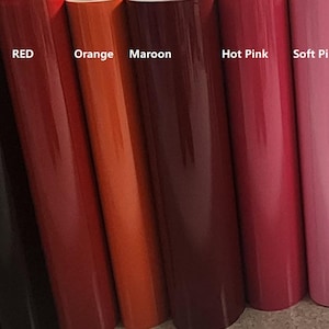 May include: A collection of vinyl rolls in various colors including Matte Flat Black, Gloss Black, Red, Orange, Maroon, Hot Pink, Soft Pink, Gold, and Silver. Each roll is labeled with its corresponding color name in white text on the surface.