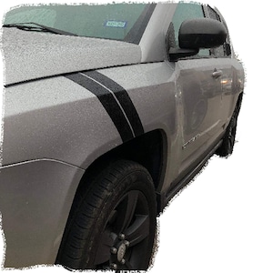 Puede incluir: Un Jeep Compass plateado con rayas laterales negras y llantas negras. El coche está aparcado en la calle.