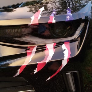 Könnte beinhalten: Ein schwarzes Auto mit einem rot-weiß-blauen amerikanischen Flaggenaufkleber auf der vorderen Stoßstange. Der Aufkleber hat die Form von Krallen.