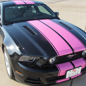 Puede incluir: Un Ford Mustang negro con rayas de carreras rosas. El coche tiene una parrilla y faros negros. El coche está aparcado en una superficie de hormigón.