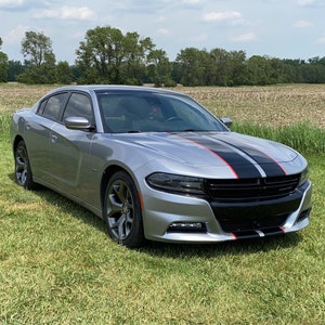 May include: A silver Dodge Charger with black and red racing stripes parked in a grassy field. The car has a blacked-out grille and tinted windows.