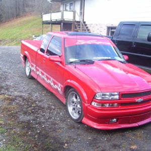 Puede incluir: Una camioneta Chevrolet S-10 roja con pintura personalizada y techo corredizo. La camioneta tiene una parrilla negra y ruedas negras. La camioneta está estacionada en un camino de grava.