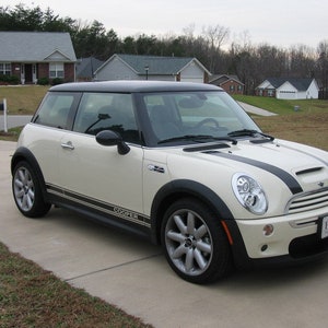 Puede incluir: Un Mini Cooper blanco con rayas de carreras negras y techo negro. El coche tiene un parachoques delantero negro y faldones laterales negros. El coche está aparcado en una entrada de coches delante de una casa.