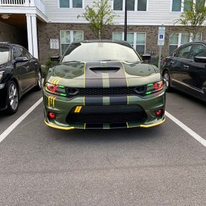 May include: A green Dodge Charger with black racing stripes and yellow accents. The car has green and black camouflage accents on the bonnet and front bumper. The car is parked in a car park.