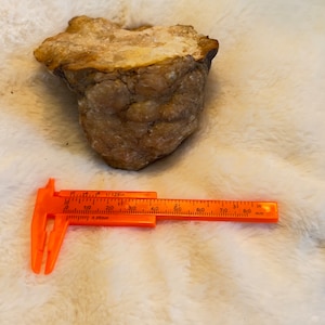 Puede incluir: Primer plano de una roca de color marrón claro, de textura rugosa, con un interior blanco y cristalino. La roca se mide con una regla, mostrando que tiene aproximadamente 6,5 cm de ancho.