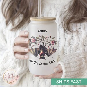 May include: A white frosted tumbler with a bamboo lid and a straw. The tumbler has a graphic of a bat hanging upside down from a branch with red flowers. The text "Ashley" is printed at the top of the tumbler and the text "Bat Out Of Hell Crazy" is printed at the bottom.