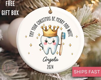 May include: White ceramic Christmas ornament with a cartoon tooth wearing a gold crown and holding a toothbrush. The ornament says "May your Christmas be merry and white" and has the name "Angela" and the year "2024" printed on it.