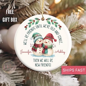 May include: A white ceramic Christmas ornament featuring two snowmen wearing hats and scarves. The ornament has the text "WE'LL BE FRIENDS UNTIL WE'RE OLD AND SENILE... THEN WE WILL BE NEW FRIENDS!" with the names Jennifer and Ashley.