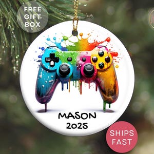 May include: A white, round ornament featuring a colorful, watercolor-style video game controller. The controller is painted in shades of blue, green, pink, and yellow. The name "MASON" and the year "2025" are printed below the controller.