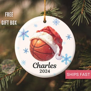 May include: White ceramic Christmas ornament with a basketball wearing a Santa hat. The basketball is brown and orange with white snowflakes in the background. The ornament is personalized with the name "Charles" and the year "2024".