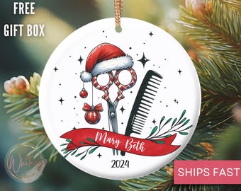 May include: A white ceramic Christmas ornament featuring a Santa hat, scissors, and a comb. A red banner reads "Mary Beth 2024". The ornament has a gold string for hanging and is decorated with stars and greenery.
