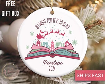 May include: White ceramic Christmas ornament with a red book open, showing a winter village scene with Santa Claus flying in a sleigh. The text "Oh what fun it is to read" is written above the book. The ornament is personalized with the name "Penelope" and the year "2024".