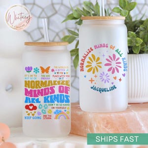 May include: Two frosted glass tumblers with bamboo lids and colorful designs. The tumblers feature the text "Normalize Minds of All Kinds" and "Don't Be So Hard on Yourself". One tumbler has the name "Jacqueline" on it.