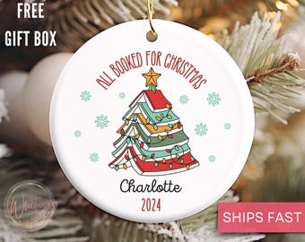 May include: White ceramic Christmas ornament with a book-shaped Christmas tree decorated with colorful lights. The text "All Booked for Christmas" is written around the tree. The ornament is personalized with the name "Charlotte" and the year "2024".