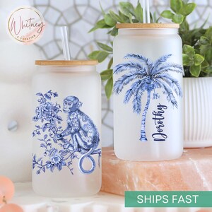 May include: Two frosted glass tumblers with bamboo lids and straws. One features a blue and white monkey design, the other a blue palm tree with the name "Dorothy". Both are on a pink surface.