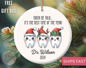 May include: White ceramic Christmas ornament with three cartoon teeth wearing Santa hats, elf hats, and reindeer antlers. The ornament says "Tooth be told... It's the best time of the year!" and "Dr. Wilson 2024".
