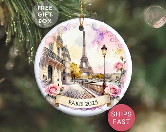 May include: A circular ceramic Christmas ornament depicting a watercolour scene of Paris, including the Eiffel Tower and streetscape. The ornament has a gold hanging loop and the text "PARIS 2025" on a banner.