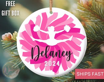 May include: Pink and white ceramic Christmas ornament with a silhouette of a cheerleader holding pom-poms. The ornament has a pink and white swirl design and the name "Delaney 2024" is written in black.