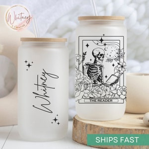 May include: Two frosted glass tumblers with bamboo lids and straws. One tumbler has a black and white illustration of a skeleton reading a book with the text "The Reader". The other tumbler has the name "Whitney" written in cursive with black stars.