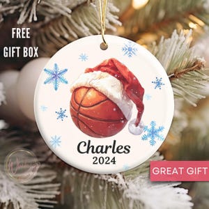 May include: White ceramic Christmas ornament with a basketball wearing a Santa hat. The basketball is orange and brown with white stitching. The ornament has blue snowflakes and the text "Charles 2024".