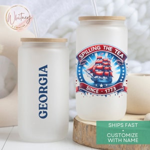 May include: Two white tumblers with lids and straws. One tumbler has the text "GEORGIA" in blue lettering. The other tumbler has a red, white, and blue graphic of a ship with the text "SPILLING THE TEA SINCE 1773".