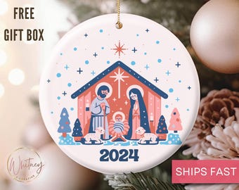 May include: A white ceramic Christmas ornament with a colorful illustration of the nativity scene. The ornament features a red and blue stable with a star on top, a yellow star in the sky, and the year 2024 printed below the stable.