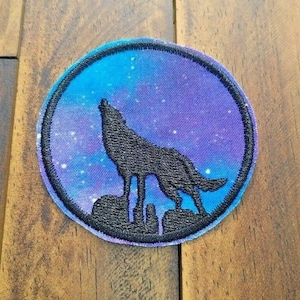 May include: A round, embroidered patch with a black silhouette of a wolf howling at the moon. The moon is a gradient of blue and purple with white stars.