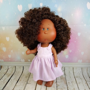 May include: A doll with dark skin, blue eyes, and a large head of brown curly hair. The doll wears a light purple dress with a textured bodice and a flared skirt. The background is a soft gradient of colors with heart shapes.