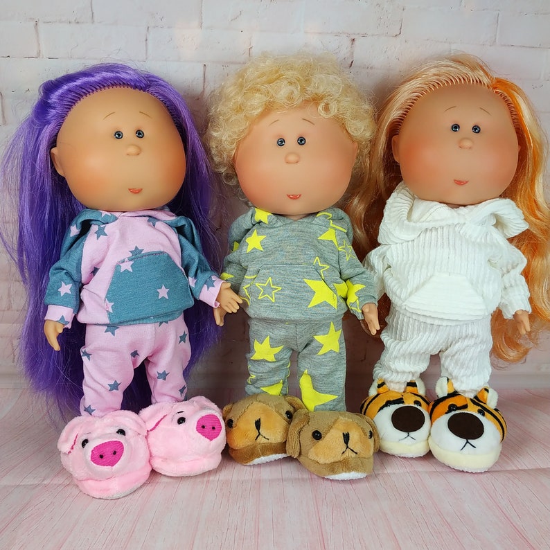 May include: Three dolls with different colored hair and clothing. The doll on the left has purple hair and is wearing a pink and blue pajamas with a star pattern. The doll in the middle has blonde hair and is wearing a gray and yellow pajamas with a star pattern. The doll on the right has orange hair and is wearing a white sweater and pants. All three dolls are wearing animal-themed slippers.