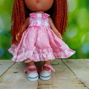 May include: A doll wearing a pink dress with white floral patterns and a gingham bodice. The dress has ruffled trim and a pink bow. The doll has red hair and is wearing pink shoes with white soles.