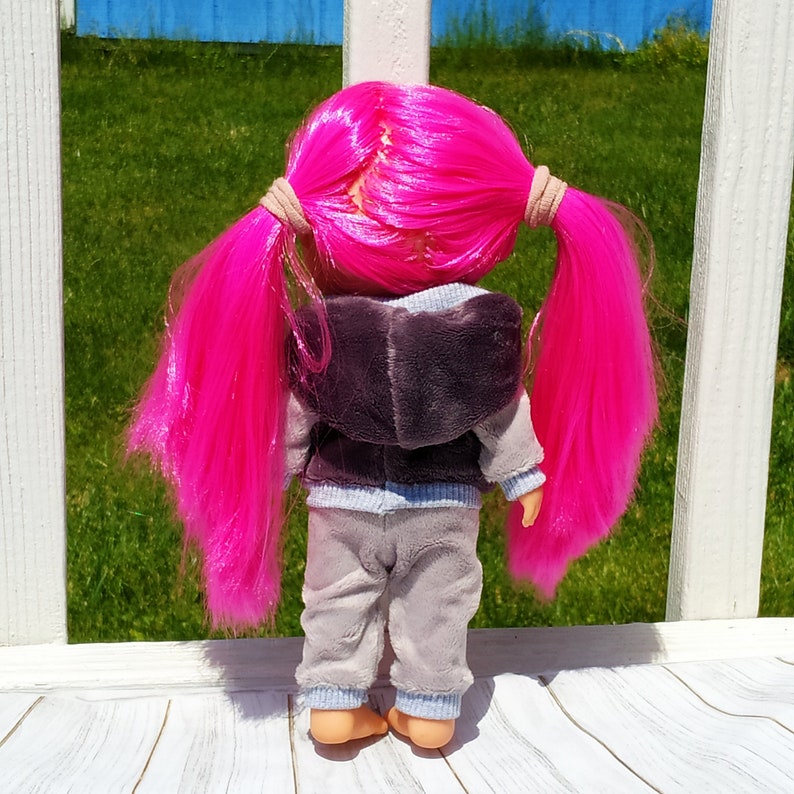 May include: A doll with long, bright pink hair in pigtails, wearing a gray hooded sweatshirt and matching sweatpants.