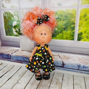 May include: A handcrafted doll with a unique appearance. The doll features a round face with blue eyes, freckles, and a large, curly, orange and pink wig. It is dressed in a black dress with colourful polka dots and matching shoes. A black bow with white polka dots is in the doll's hair.