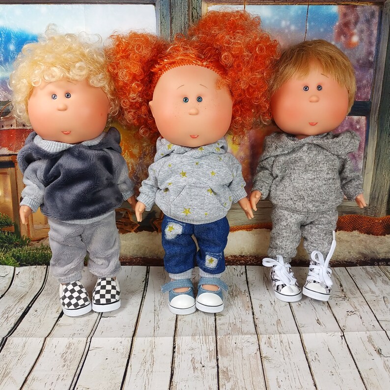 May include: Three handmade dolls with different colored hair and clothing. The dolls are wearing grey hoodies and pants, and one doll is wearing blue jeans. The dolls are standing on a wooden surface.
