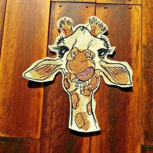 May include: Embroidered patch of a giraffe's face with large eyes and a tongue sticking out. The giraffe is brown and white with a beige background.