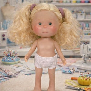 Może przedstawiać: Ręcznie robiona lalka z blond kręconymi włosami w kucykach, ubrana w białe dzianinowe szorty. Lalka ma różowe policzki i zielone oczy. Tło przedstawia materiały do szycia, sugerując motyw rzemieślniczy lub zabawkarski.