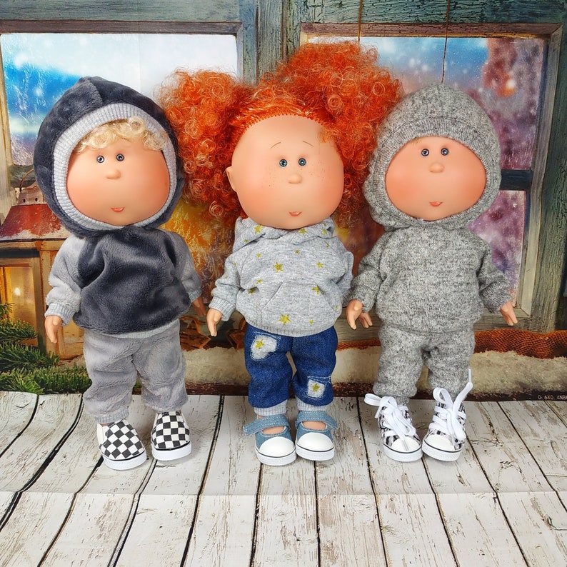May include: Three handmade dolls wearing hooded sweatshirts and jeans. The dolls have rosy cheeks and large eyes. The dolls are standing on a white wooden surface.