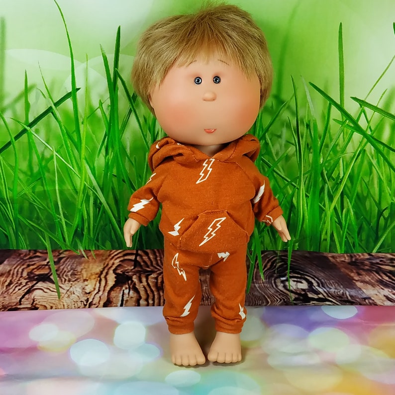 May include: A handmade doll with a round face, blonde hair, and large blue eyes. The doll is wearing a brown hoodie and matching pants with a white lightning bolt pattern.