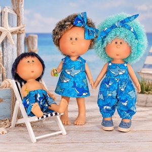 May include: Three dolls with round faces and freckles. One doll with dark hair sits in a miniature striped beach chair wearing shark-print shorts. A second doll with brown curly hair wears a shark-print dress and bow. A third doll with bright blue curly hair wears a shark-print romper and headband.