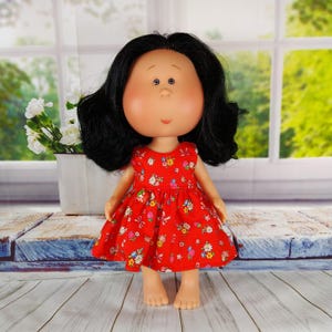 May include: A doll with black hair and a red dress decorated with a floral pattern. The doll has a round face and is standing on a wooden surface. The dress features a variety of colourful flowers.