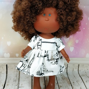 May include: A doll with brown curly hair wearing a white dress with a black Eiffel Tower print. The doll has brown skin and blue eyes.