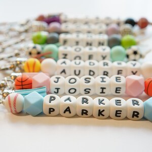 Pu&ograve; includere: Portachiavi con nomi scritti con perline di lettere bianche. I portachiavi presentano varie perline colorate e ciondoli a tema sportivo, tra cui palline da baseball, basket e calcio. I nomi "PARKER", "JOSIE", "AUDREY" e "ZOEY" sono visibili.