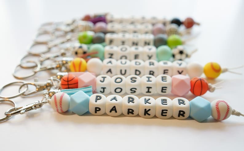 Pu&ograve; includere: Portachiavi con nomi scritti con perline di lettere bianche. I portachiavi presentano varie perline colorate e ciondoli a tema sportivo, tra cui palline da baseball, basket e calcio. I nomi "PARKER", "JOSIE", "AUDREY" e "ZOEY" sono visibili.