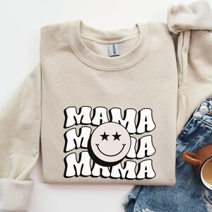 May include: A beige sweatshirt with a black and white graphic design featuring the word "MAMA" repeated three times with a smiley face in the middle. The smiley face has two black stars for eyes.