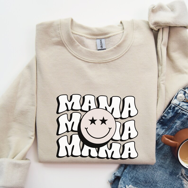 May include: A beige sweatshirt with a black and white graphic design featuring the word "MAMA" repeated three times with a smiley face in the middle. The smiley face has two black stars for eyes.