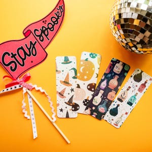 Könnte beinhalten: Eine Sammlung von Lesezeichen zum Thema Halloween mit Hexenhüten, Kürbissen und Zaubertränken. Ein rosa Banner mit der Aufschrift "Stay Spooky" und eine Discokugel sind ebenfalls im Bild. Die Artikel sind auf einer orangefarbenen Oberfläche angeordnet.