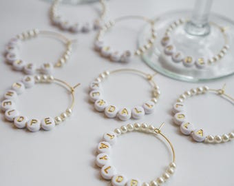 May include: Several gold hoop wine charms with white letter beads spelling out names. The charms are accented with small pearls. The names include Lola, Taylor, Kendall, and Hope. The charms are on a white surface.