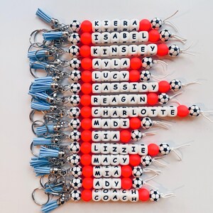 Pu&ograve; includere: Una collezione di portachiavi personalizzati con nomi scritti con perline di lettere bianche. Ogni portachiavi presenta perline rosse, perline a forma di pallone da calcio e un nappa blu. I nomi includono Kiera, Isabel, Kinsley, Ayla, Lucy, Cassidy, Reagan, Charlotte, Madi, Gia, Lizzie, Macy, Mia, Ady e Coach.