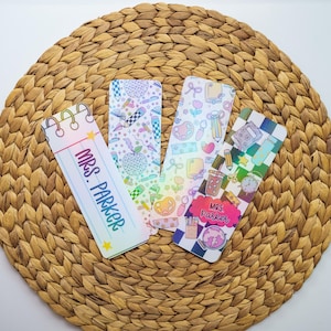 以下が含まれることがあります： さまざまなデザインの、教師をテーマにしたカラフルなブックマークが4つ。1つのブックマークは「MRS. PARKER」というテキストが入ったメモ帳のデザインです。他のブックマークには、鉛筆、リンゴ、パレットなどの学校関連のグラフィックがあります。ブックマークは、織られた円形の表面にあります。